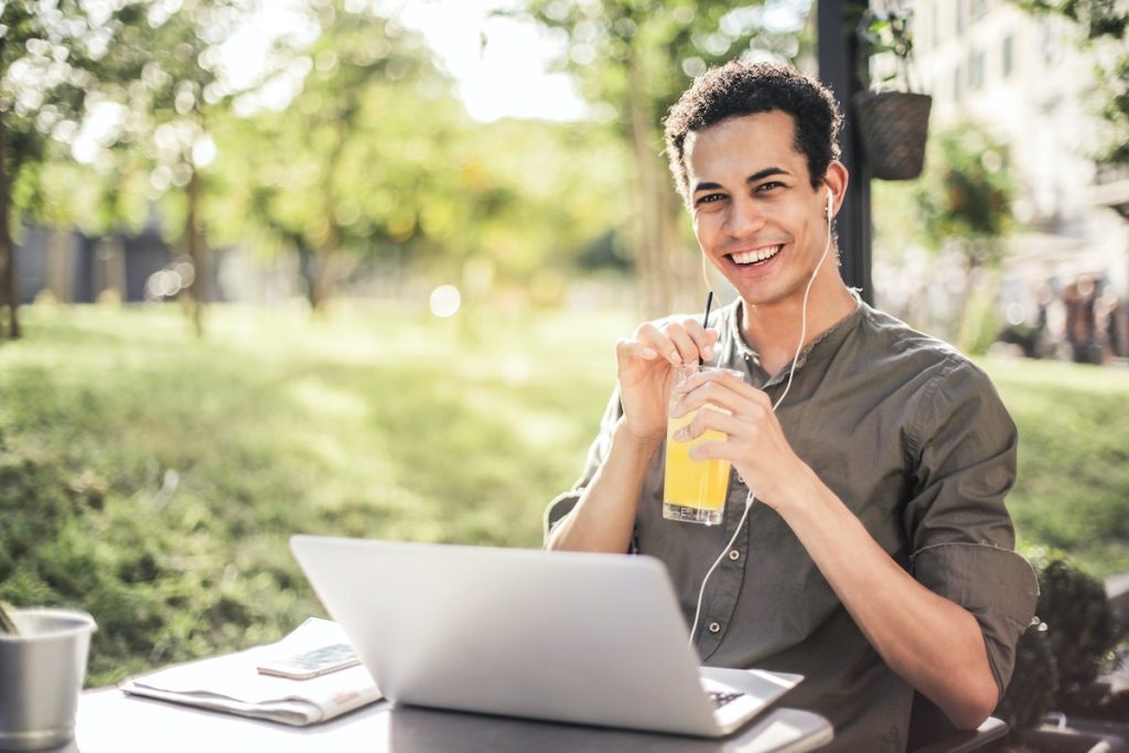 Un joven sentado en una mesa al aire libre con una computadora portátil y un jugo de naranja, encontrando refugio a través de la música contemplativa.